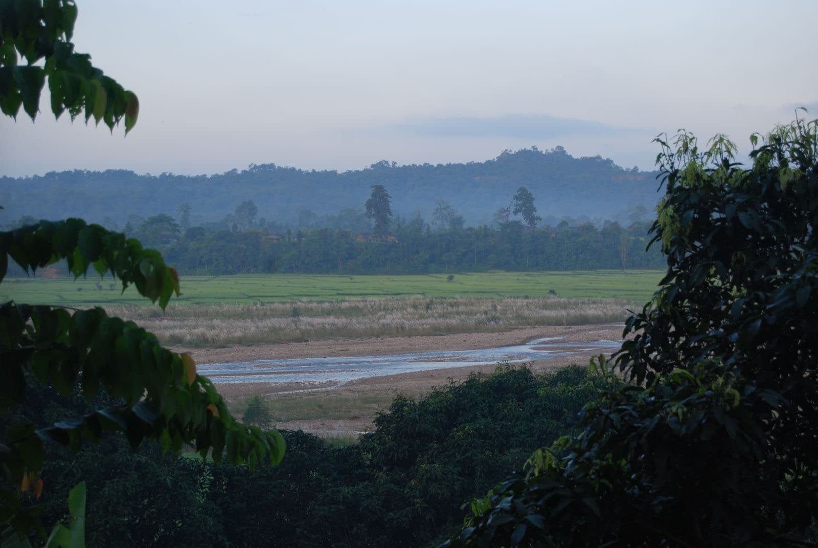 Green fields and a river bed below forested hills at dusk in Nepal's Terai